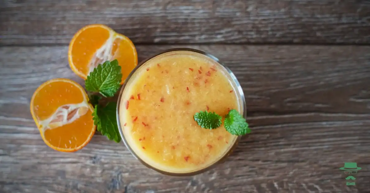 A glass of orange smoothie with mint leaves on top, surrounded by halved oranges and more mint, on a wooden surface.