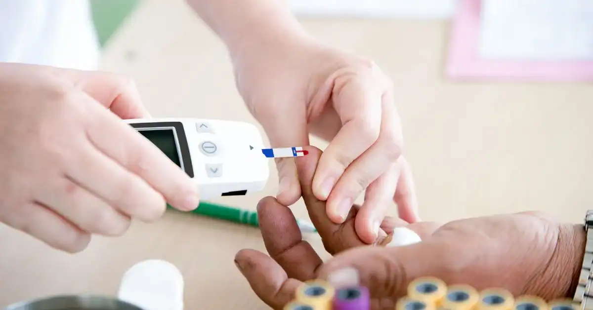 A close-up of a person's hand being tested for blood sugar with a glucose meter, held by another person, with test strips and vials in the background on a table.