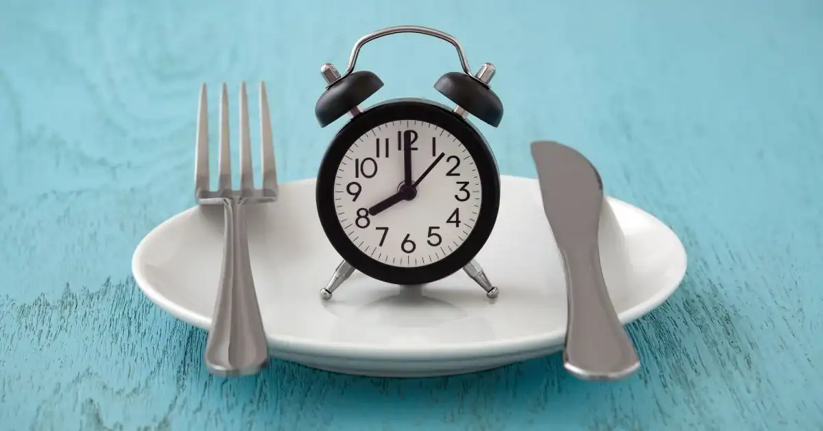 A white plate with a black alarm clock showing 10:47, flanked by a fork and knife, placed on a blue wooden surface.