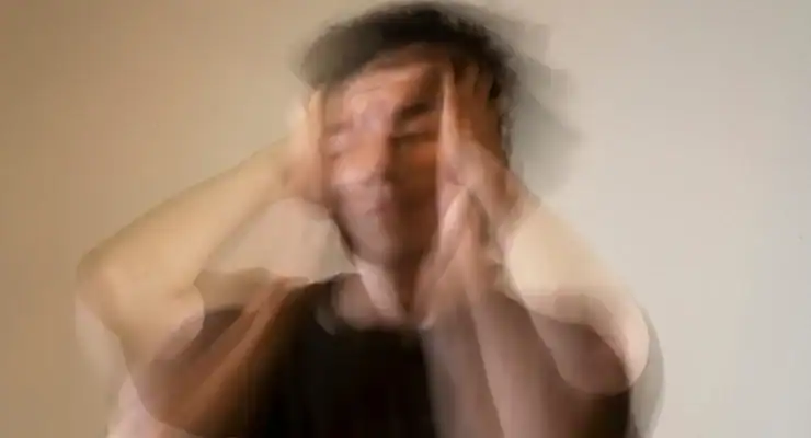 A blurry image of a person with hands on their head, appearing distressed, against a plain background.