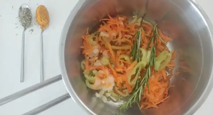 A metal bowl with shredded carrots, onion, garlic, peppers, and rosemary. Two spoons with turmeric and dill are beside it on a white surface.